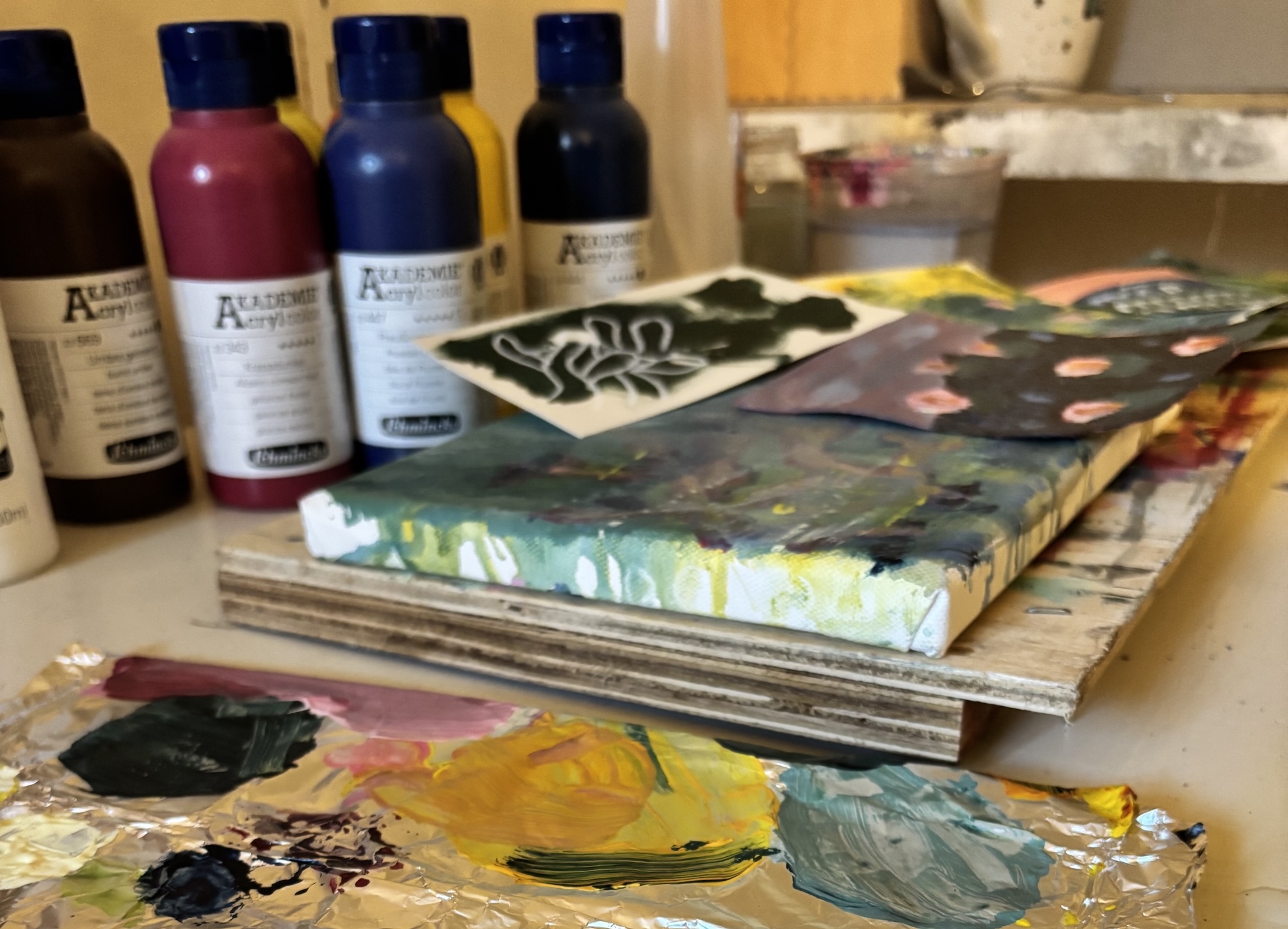 Atelierfoto: Acrylfarben, eine bemalte Leinwand und eine Palette aus Alufolie mit angemischten Farbtupfern